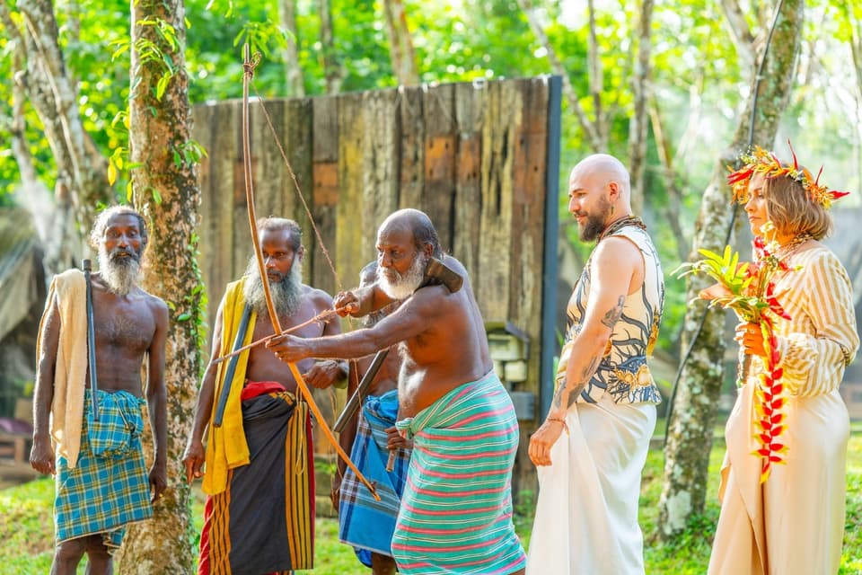 A community elder demonstrating the traditional bow as the couple watches in the daylight forest