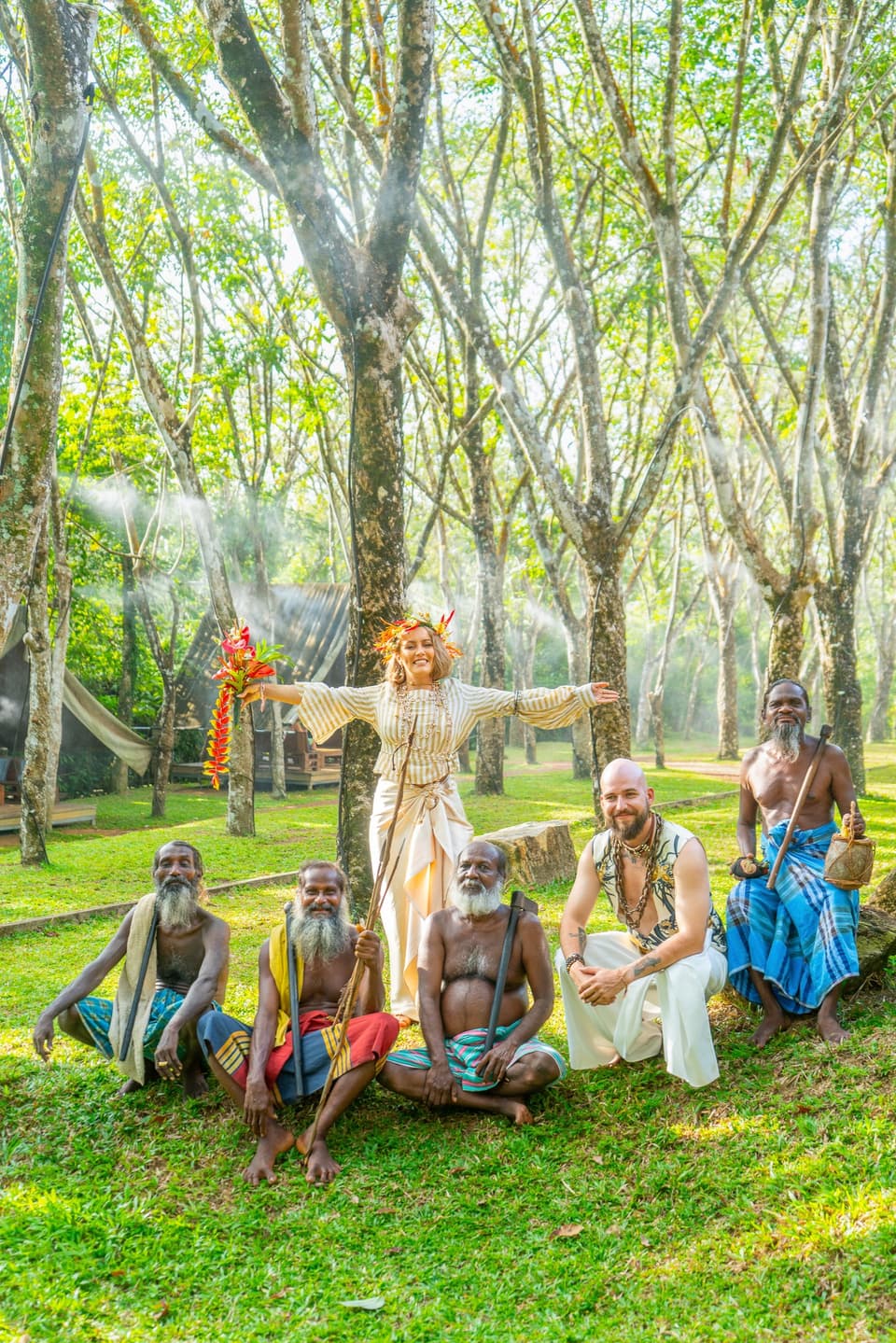 Bride with open arms posing alongside the groom and Vedda elders on a sunlit forest lawn