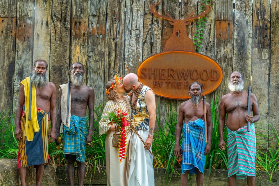 Couple sharing a kiss before the Sherwood sign, flanked by Vedda community elders